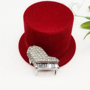 May include: A silver-toned brooch shaped like a grand piano, encrusted with clear rhinestones. The piano keys are made of clear baguette-shaped rhinestones.