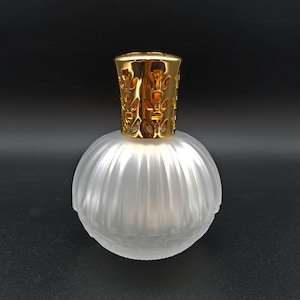 May include: A frosted glass diffuser bottle with a gold metal lid. The bottle has a round shape with a textured surface.