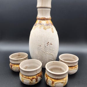 May include: A ceramic sake set featuring a bottle and four small cups. The bottle is off-white with a textured surface and a brown and gold band near the top. The cups have a similar design, with a brown and gold band around the base.