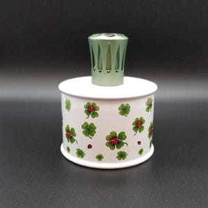 May include: A cylindrical white fragrance lamp with a repeating pattern of green four-leaf clovers and small red ladybugs. The top of the lamp features a metallic light green vented cap with vertical cutouts, centered on the flat white lid.