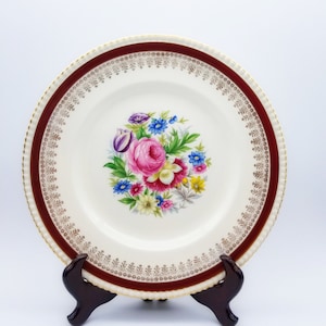 Puede incluir: Un plato de cena de cerámica blanca con un borde burdeos y un borde dorado. El centro del plato presenta un diseño floral con flores rosas, rojas, amarillas y azules.