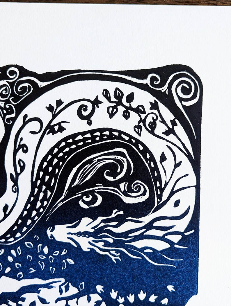 May include: Black and white linocut print of a dragon with blue ink. The dragon is curled up with its head and eyes visible. The background is a dark blue colour with white details.