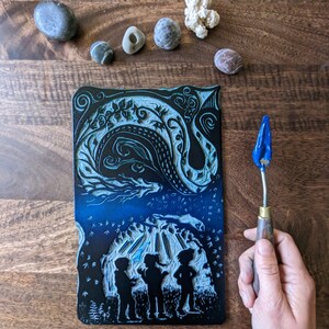 May include: A black and blue linoleum block print depicting three figures standing in front of a cave with a swirling design above. The print is on a wooden surface with a blue and silver tool for carving linoleum.