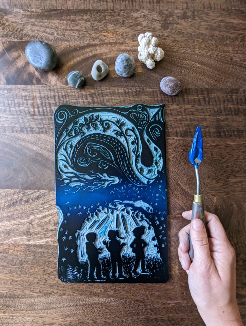 May include: A black and blue linoleum block print depicting three figures standing in front of a cave with a swirling design above. The print is on a wooden surface with a blue and silver tool for carving linoleum.