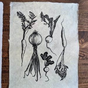 May include: A rectangular piece of off-white, textured handmade paper featuring a black ink block print illustration of five different root vegetables, including carrots, an onion, and radishes with their leafy green tops.