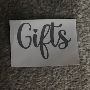 May include: A rectangular sticker with the word "Gifts" in a cursive font. The word is in a dark gray color, and a small heart is above the letter "i". The sticker is on a white background.