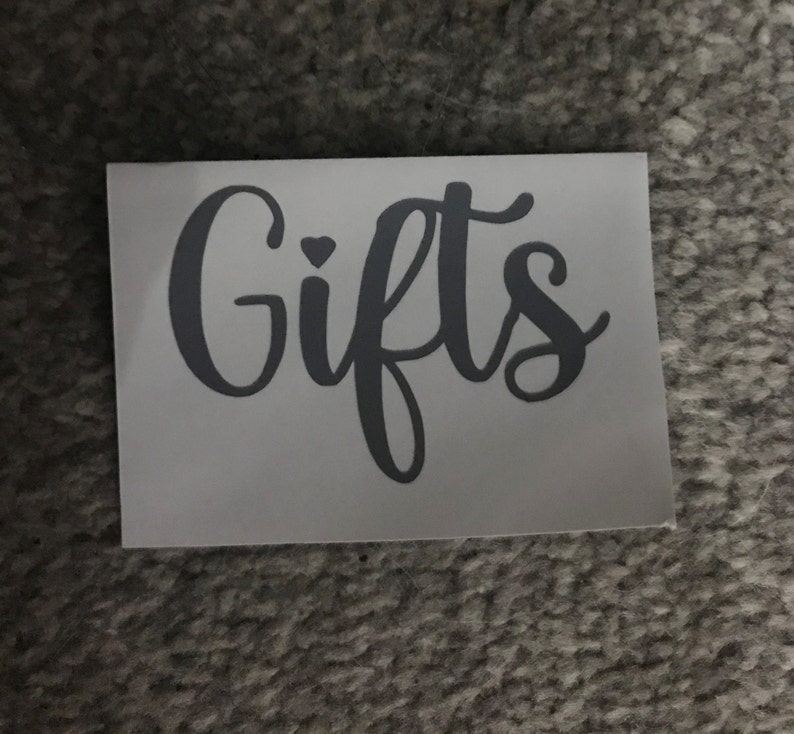 May include: A rectangular sticker with the word "Gifts" in a cursive font. The word is in a dark gray color, and a small heart is above the letter "i". The sticker is on a white background.