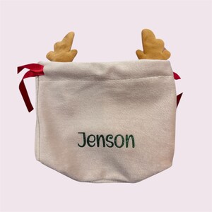 May include: A light beige drawstring bag embroidered with the name "Jenson" in green. The bag features two small, tan reindeer antlers and red ribbon ties, perfect for gifting.