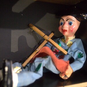 May include: A marionette puppet with a painted face, blue eyes, and red lips, wearing a light blue outfit decorated with colorful sequins. The puppet features a black hat and is attached to a wooden cross-shaped control bar with black strings.