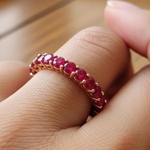 May include: A rose gold ring set with vibrant red gemstones. The ring is worn on a finger, showcasing the polished metal and the rich color of the stones. The ring's design features a continuous row of gemstones.