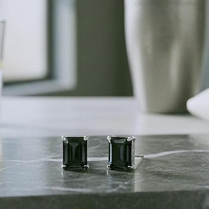 May include: A pair of black emerald-cut stud earrings with silver settings. The earrings are on a grey marble tray, next to a cocktail shaker and a glass of water with a lemon slice.