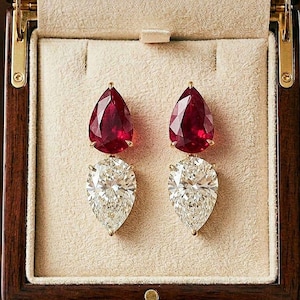 May include: A pair of elegant drop earrings displayed in a wooden jewelry box. Each earring features a pear-shaped, vibrant red gemstone above a pear-shaped, sparkling white diamond. The earrings are set in gold and presented against a cream-colored backdrop.