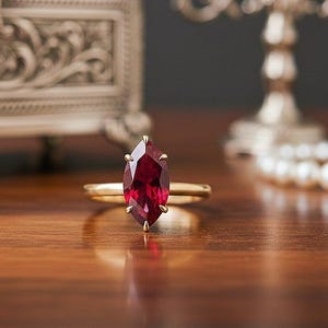 May include: A gold engagement ring featuring a marquise-cut ruby. The ring is the central element, set against a blurred backdrop including a decorative jewellery box, a candelabra, and a pearl necklace. The ruby is a deep red colour.