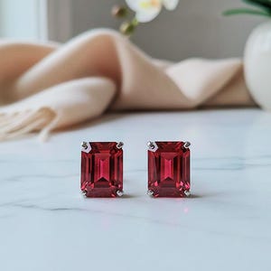 May include: Two silver stud earrings, each with a rectangular, deep red gemstone. The earrings are presented on a white marble surface, with a cream-coloured scarf and a vase of white orchids in the background.