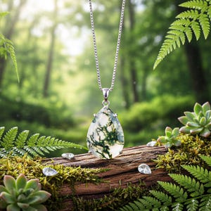 Puede incluir: Un collar de plata con un colgante en forma de lágrima. El colgante presenta una piedra translúcida con inclusiones verdes en forma de helecho. El collar se exhibe sobre un tronco cubierto de musgo con pequeñas gemas claras y follaje verde en el fondo.