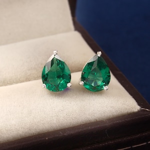 May include: A pair of silver stud earrings featuring large, pear-shaped, faceted emerald green gemstones. The earrings are set in a silver metal and are displayed in a dark brown jewelry box with a light beige interior.