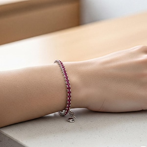 May include: A silver bracelet featuring a line of small, bright red gemstones. The bracelet is delicate and appears to be made of silver. The gemstones are closely set, creating a continuous line of colour. The bracelet is on a wrist.