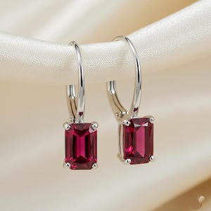 May include: A pair of silver-toned leverback earrings, each featuring a rectangular, deep red gemstone. The earrings are displayed against a soft, off-white fabric background, highlighting the jewelry's design and color.