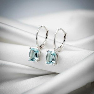 Puede incluir: Un par de pendientes de plata con cierre de palanca y piedras preciosas rectangulares de color azul claro. Los pendientes se exhiben sobre una tela blanca y sedosa, creando una presentación suave y elegante. Las piedras miden aproximadamente 1,3 cm de largo.