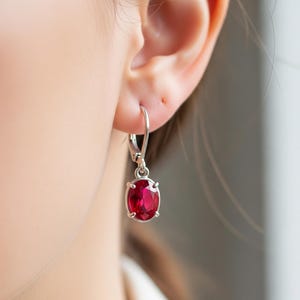 May include: A close-up of a silver earring with a vibrant red oval gemstone. The earring is suspended from a lever back, showcasing the polished gemstone's rich color and reflective qualities. The jewelry is set against a blurred background.