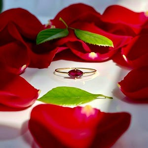 May include: A gold ring with a marquise-cut ruby gemstone, accented with a small diamond. The ring is set against a backdrop of red rose petals and green leaves, creating a romantic and elegant aesthetic.