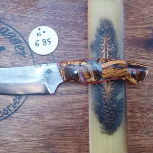 Peut inclure: Un couteau artisanal avec un manche en bois brun et rouge et une lame argentée. Le couteau est posé sur une surface en bois avec un tampon circulaire qui indique "Raven's Dagger" et une étiquette blanche avec le numéro "685".