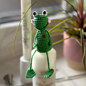 Könnte beinhalten: Eine handgefertigte grüne Häkelfrosch mit schwarz-weißen Augen, rosa Wangen und einem roten Herz. Der Frosch sitzt auf einer weißen Kerze mit Text. Eine weiße Kerze und Grünzeug sind im Hintergrund.