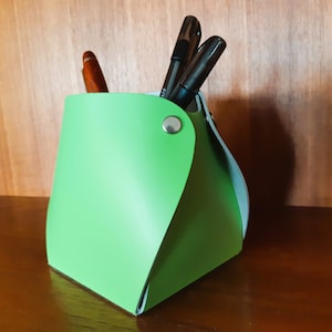 Peut inclure: Un organiseur de bureau en cuir vert de forme triangulaire avec un bouton argenté. L'organisateur contient des stylos et des crayons.