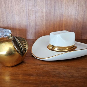 May include: A gold-coloured apple-shaped ornament with a bronze leaf sits beside a white cowboy hat-shaped dish with a gold band. The hat has a white top and gold trim.