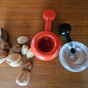May include: Two nutcrackers, one red and one clear plastic, with a black handle, on a wooden surface with almonds and hazelnuts.