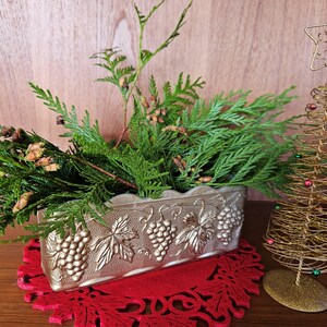 Peut inclure: Un pot en argent avec un motif de vigne de raisin contient un bouquet de branches de cèdre vert. Le pot est posé sur une dentelle rouge. Un sapin de Noël en fil de fer doré avec des perles rouges et vertes est en arrière-plan.