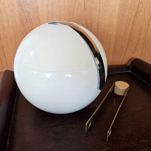 May include: A white spherical ice bucket with a silver lid and a pair of silver tongs. The ice bucket is sitting on a dark brown surface.