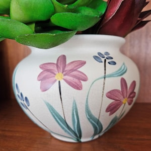 May include: A white ceramic vase with a floral design. The vase features pink and blue flowers painted on it with green leaves. The vase is sitting on a wooden surface.