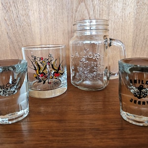 May include: Four clear glass drinking glasses with Sailor Jerry branding. Two shot glasses and a larger glass with a nautical design. A clear glass mason jar with a handle and Sailor Jerry branding.