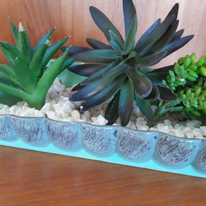 Peut inclure: Un pot en céramique turquoise avec un bord festonné, rempli de galets blancs et de trois types de succulentes. Les succulentes sont vertes et ont des textures variées.