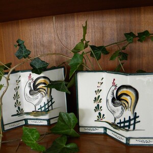 May include: Two white ceramic decorative plates with a rooster design. Each plate features a rooster illustration in black, yellow, and white, with a small fence and green accents. The plates are displayed on a wooden surface.
