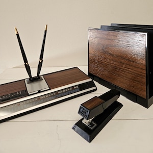May include: A retro office desk set including a pen holder with two black pens, a calendar, a stapler, and a letter holder. The set has a wood grain finish with black detailing. The stapler reads "BATES 325".