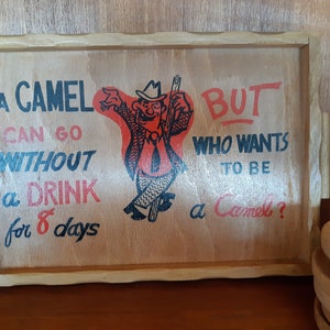 Peut inclure: Un plateau en bois avec une illustration humoristique d'un homme portant un chapeau et une cravate. Le texte sur le plateau est : "Un chameau peut se passer de boisson pendant 8 jours MAIS qui veut être un chameau ?"