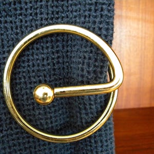 Peut inclure: Gros plan sur une boucle en métal doré avec un design circulaire et un petit accent en forme de boule. La boucle est placée sur un tissu bleu foncé texturé, peut-être un sac à main ou une ceinture.