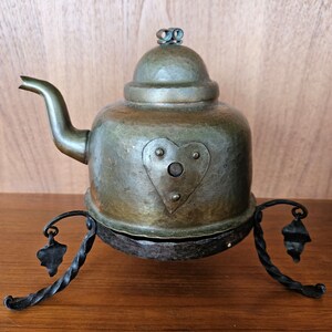 May include: An antique copper teapot with a heart-shaped design and a black metal stand. The teapot has a hammered finish and a small knob on the lid.