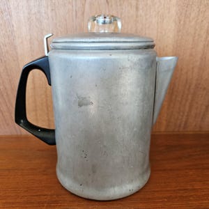 May include: A vintage aluminium coffee pot with a black handle and a clear glass knob on the lid. The pot has a spout for pouring and is on a wooden surface. A classic kitchenware item.