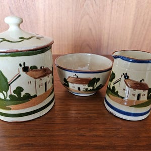 May include: A set of three ceramic kitchenware pieces with a cottagecore design. The set includes a lidded jar, a bowl, and a jug. Each piece features a white cottage with a brown roof and green trees, painted on a cream background with brown, green, and blue accents.