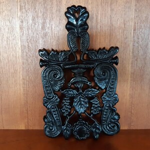 May include: Black cast iron hot pad with an ornate design featuring grapes and leaves.