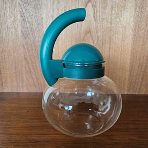 May include: A teal glass teapot with a teal plastic lid and handle. The teapot is sitting on a wooden surface.