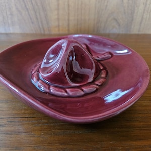 May include: A burgundy ceramic sombrero ashtray. The sombrero is detailed with a rope trim.