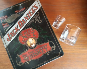 May include: A black metal tin with a red and gold label that reads "Jack Daniel's Old No. 7 Old Time Tennessee Whiskey" and "Lem Motlow Prop.". The tin is next to two clear glass shot glasses with the Jack Daniel's logo.