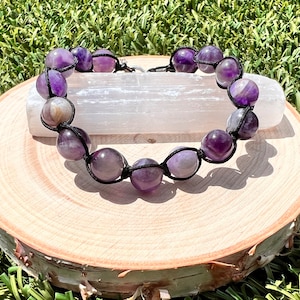 May include: A purple amethyst gemstone bracelet with black cord and beads. The bracelet is displayed on a wooden surface.