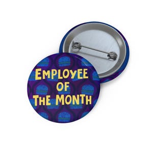May include: Two round pinback buttons. One button features the text "EMPLOYEE OF THE MONTH" in yellow, with a blue and purple burger pattern. The other button is silver with a blue rim.