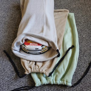 May include: Three fleece helmet bags in beige, tan, and light green. The beige bag holds a helmet with "MOTUL" and "USHIBATAI" text. Each bag has a drawstring closure with black cords. The bags are on a gray carpet.