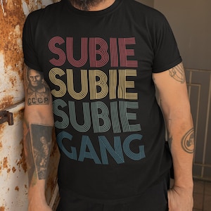 以下が含まれることがあります： レトロなスタイルのグラフィックがプリントされた黒いTシャツ。カラフルな重ねられたテキストで「SUBIE SUBIE SUBIE GANG」と書いてあります。
